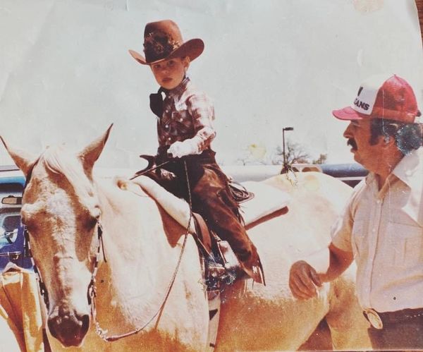 Young boy in cowboy attire rides a white horse while a man stands nearby.