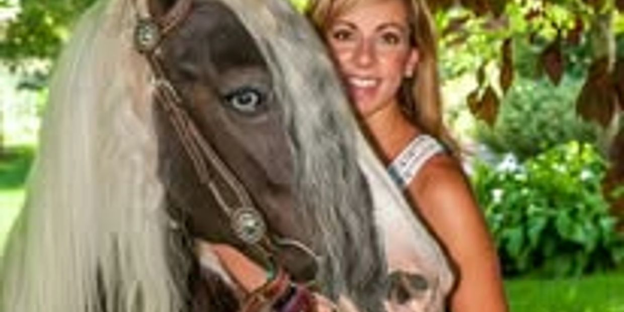 Woman smiling with a black and white horse under green foliage.