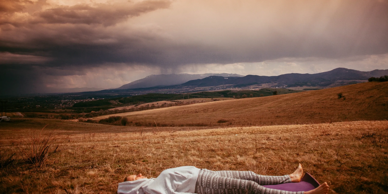 A person is lying in a field, face up to the sky. There are distant mountains under a cloudy sky.  