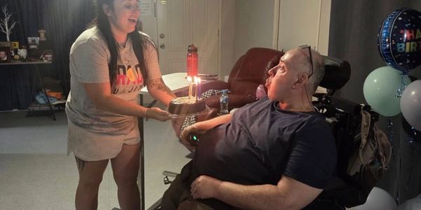 A woman presents a birthday cake with candles to a man in a wheelchair.