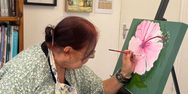 Elderly woman painting a large pink hibiscus flower on a canvas.