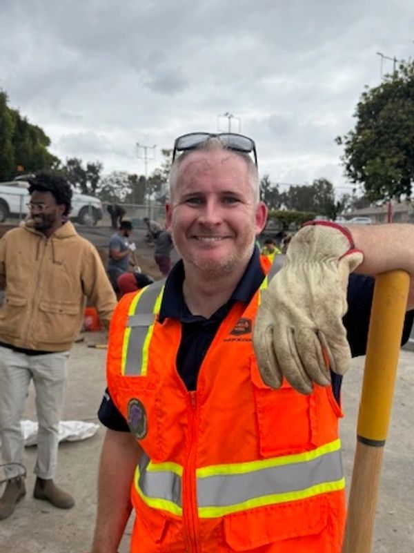 Smiling construction worker in orange safety vest holding a shovel and gloves.