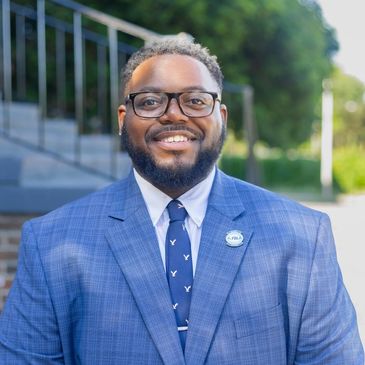 A smiling man in a blue plaid suit and glasses outdoors.