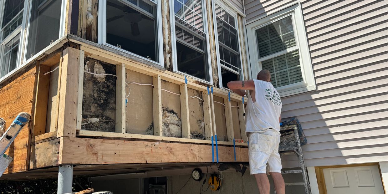 Worker repairing house exterior with exposed wooden frame and windows.