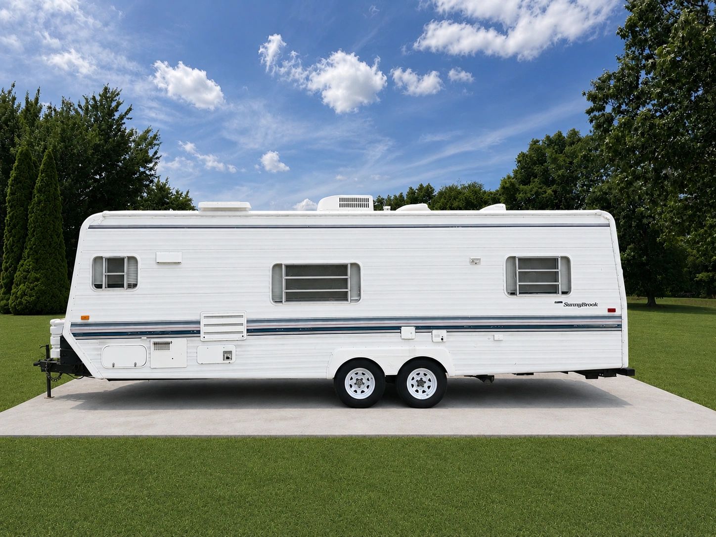 White SunnyBrook travel trailer parked on a concrete pad with trees and blue sky in the background.