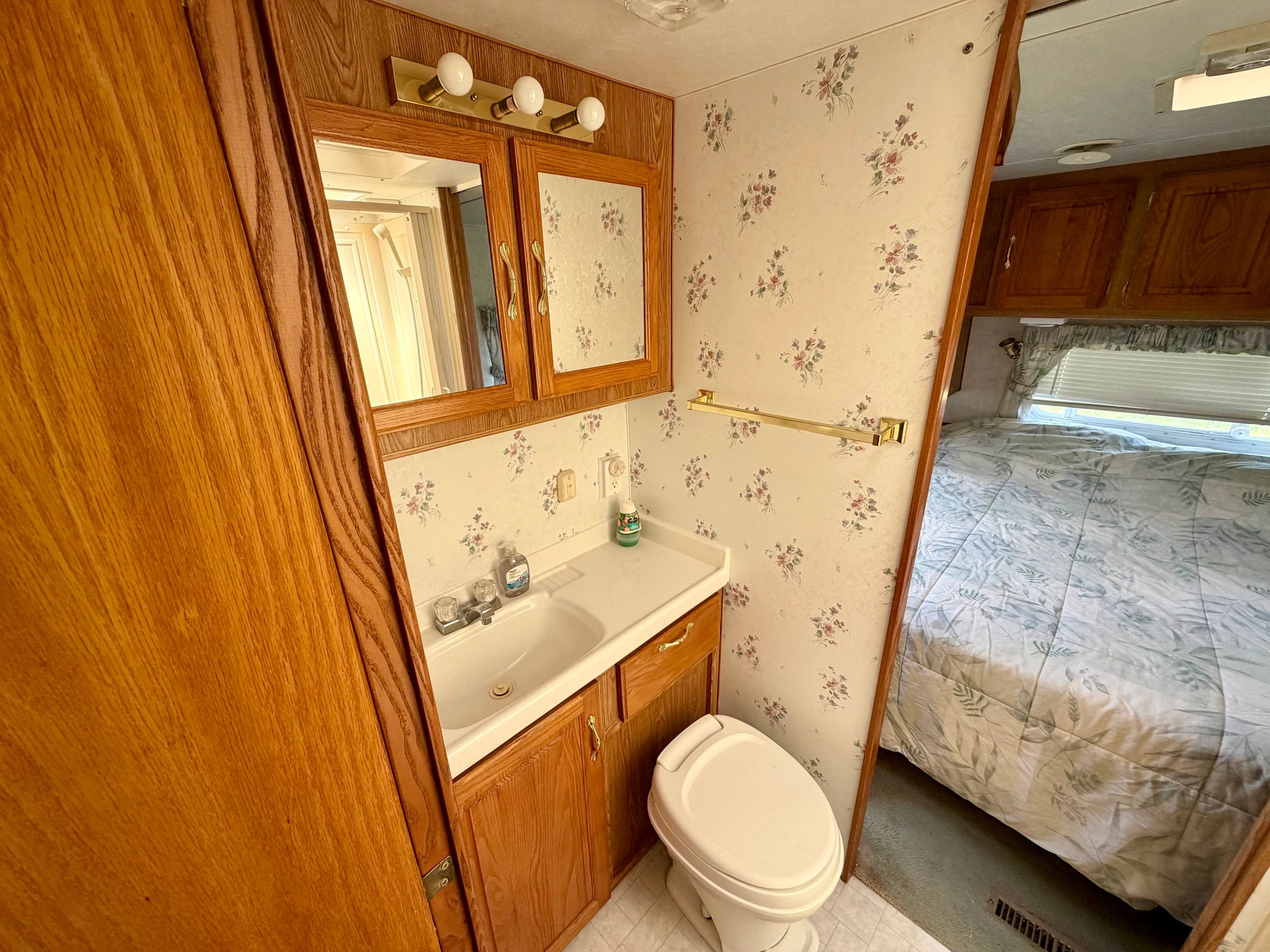 Compact bathroom with wooden cabinets and floral wallpaper in an RV or trailer.