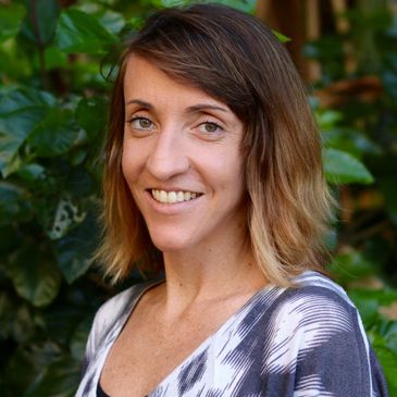Smiling woman with shoulder-length hair standing outdoors with greenery in the background.