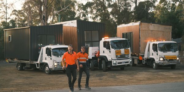 Workers in orange shirts walk past trucks carrying modular homes in a wooded area.