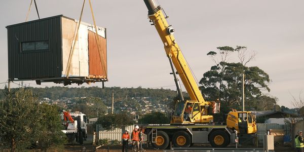 Crane lifting a small modular house at a construction site with workers around.