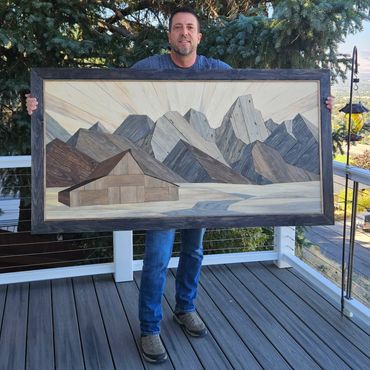 Man proudly holding framed mountain landscape artwork outdoors.