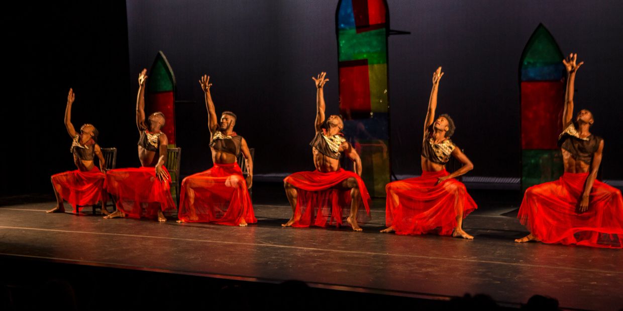 Six male dancers in red skirts perform a synchronized, expressive dance on stage.