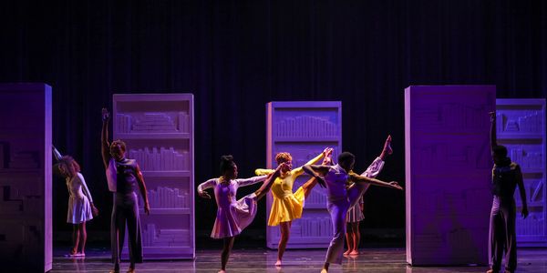 Dancers perform gracefully on stage with purple-lit bookshelves as backdrop.
