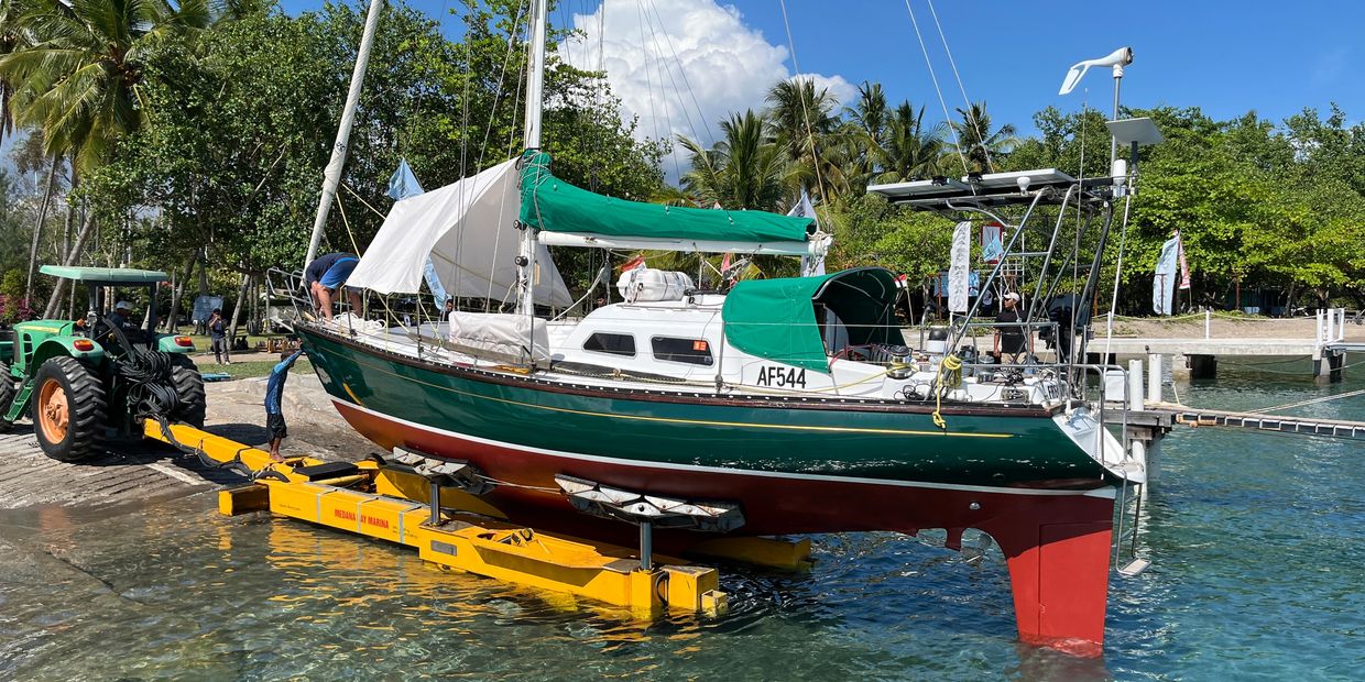 Sailboat being launched into water by tractor near palm trees.