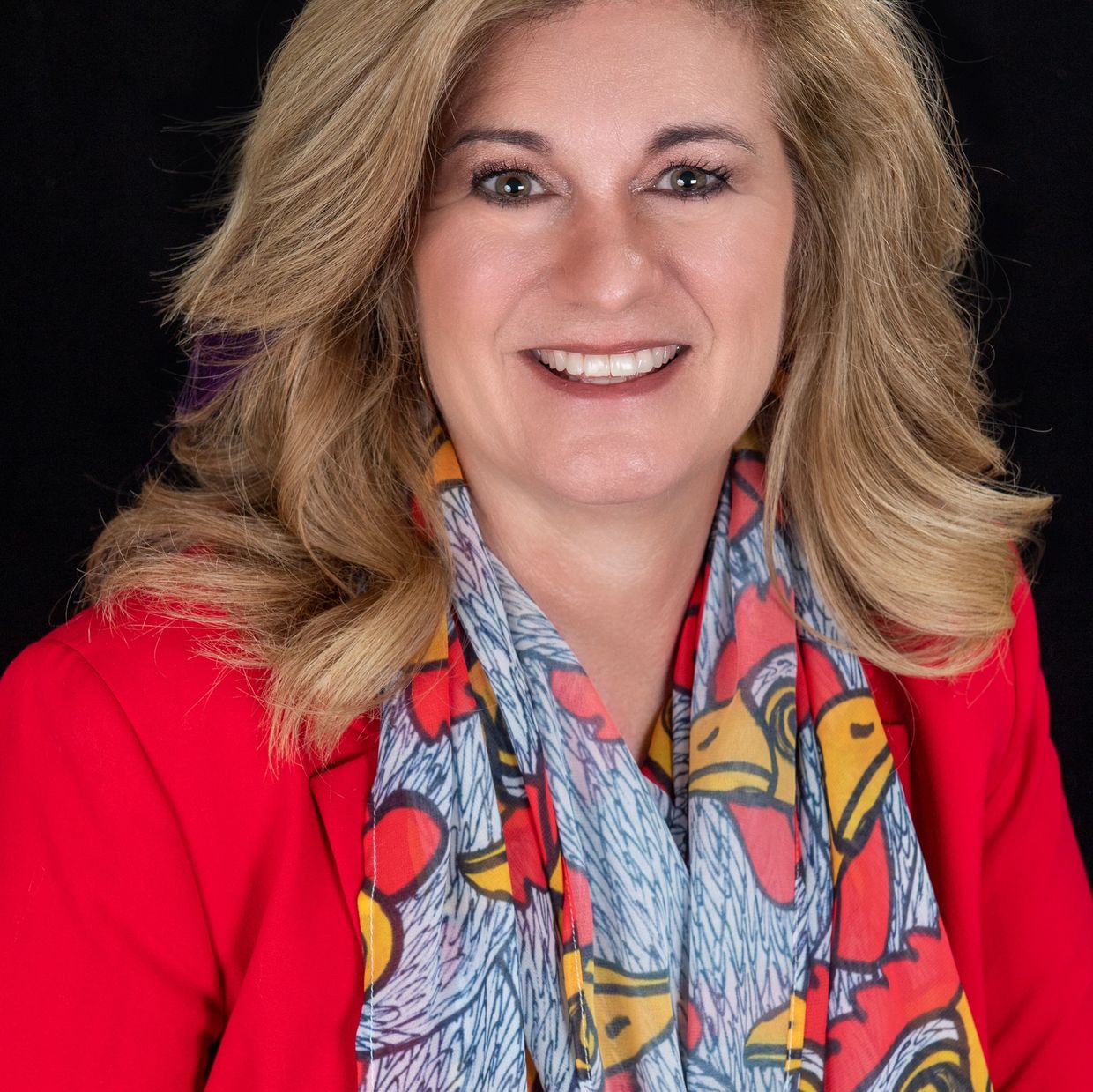 Smiling woman with blonde hair, wearing a red blazer and a colorful scarf with rooster designs.