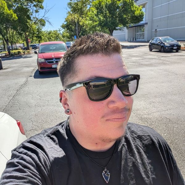Man wearing sunglasses and black shirt in a sunny parking lot.