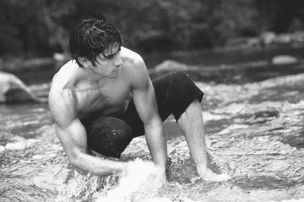 A muscular man crouches in a shallow stream, splashing water with his hands.
