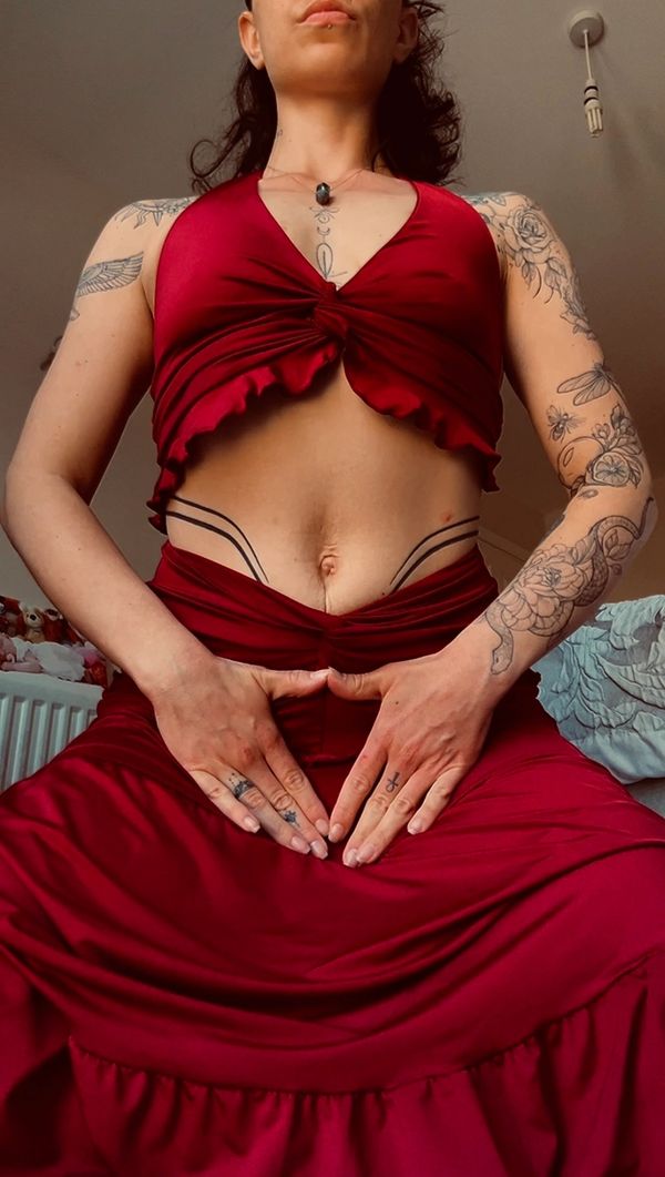 Person in red outfit with tattoos on arms and torso, hands forming a heart shape near the waist.