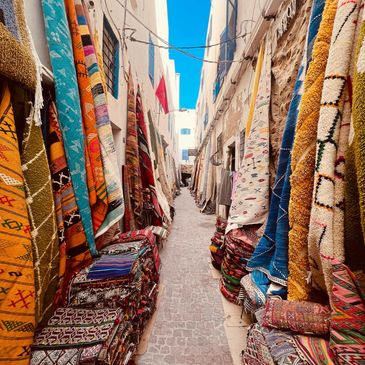 Narrow alley lined with colorful traditional rugs and carpets under a bright blue sky.