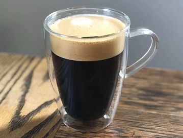 A clear glass cup filled with black coffee topped with froth.