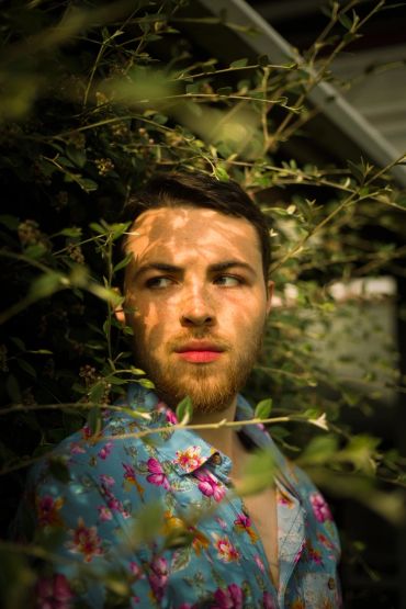 Man with floral shirt peering through green foliage.