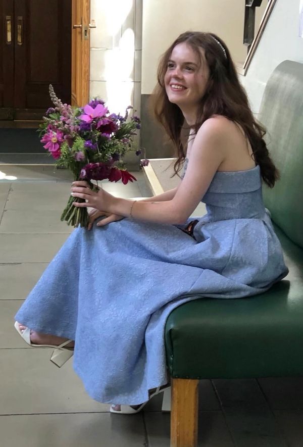 Young woman in a blue dress holding a bouquet, sitting on a bench.