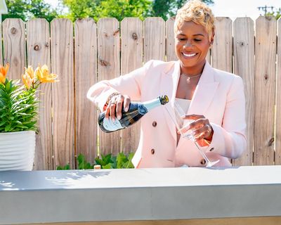Woman in pink blazer pouring champagne outdoors.