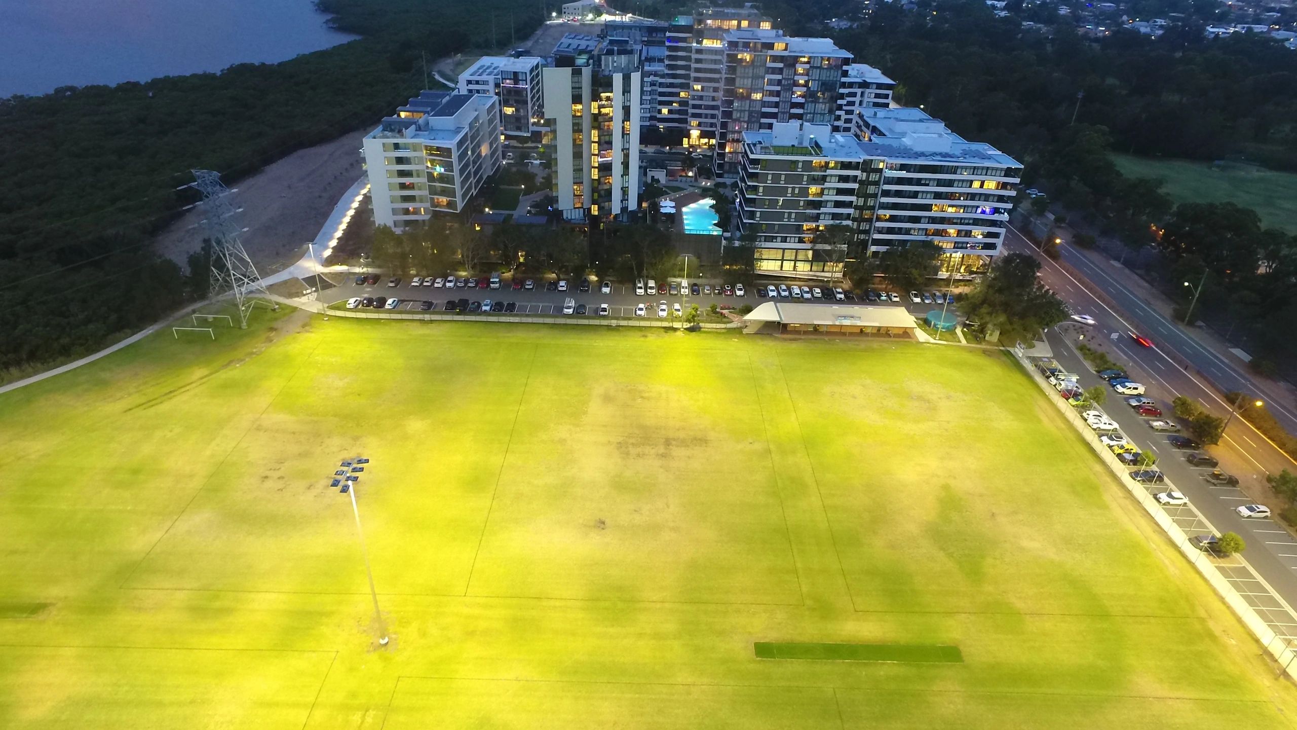 A lit sports field with modern buildings in the background at dusk.