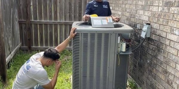 Two men inspecting an outdoor air conditioning unit beside a brick wall.