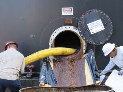 Workers manage sludge removal from an industrial tank with safety gear.