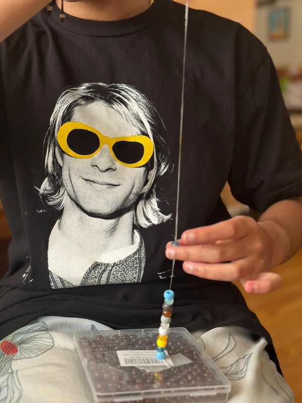 Person wearing a black Nirvana shirt with yellow sunglasses, threading beads.