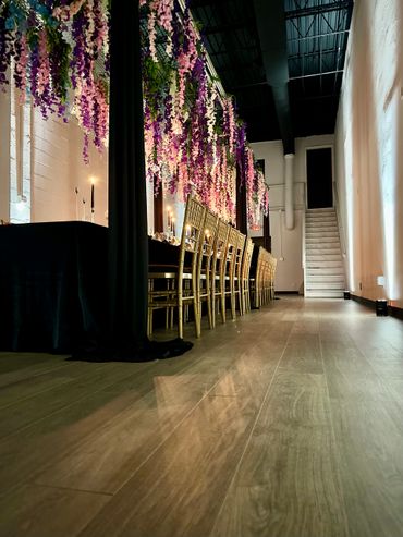 Elegant dining setup with hanging purple flowers and gold chairs in a modern venue.