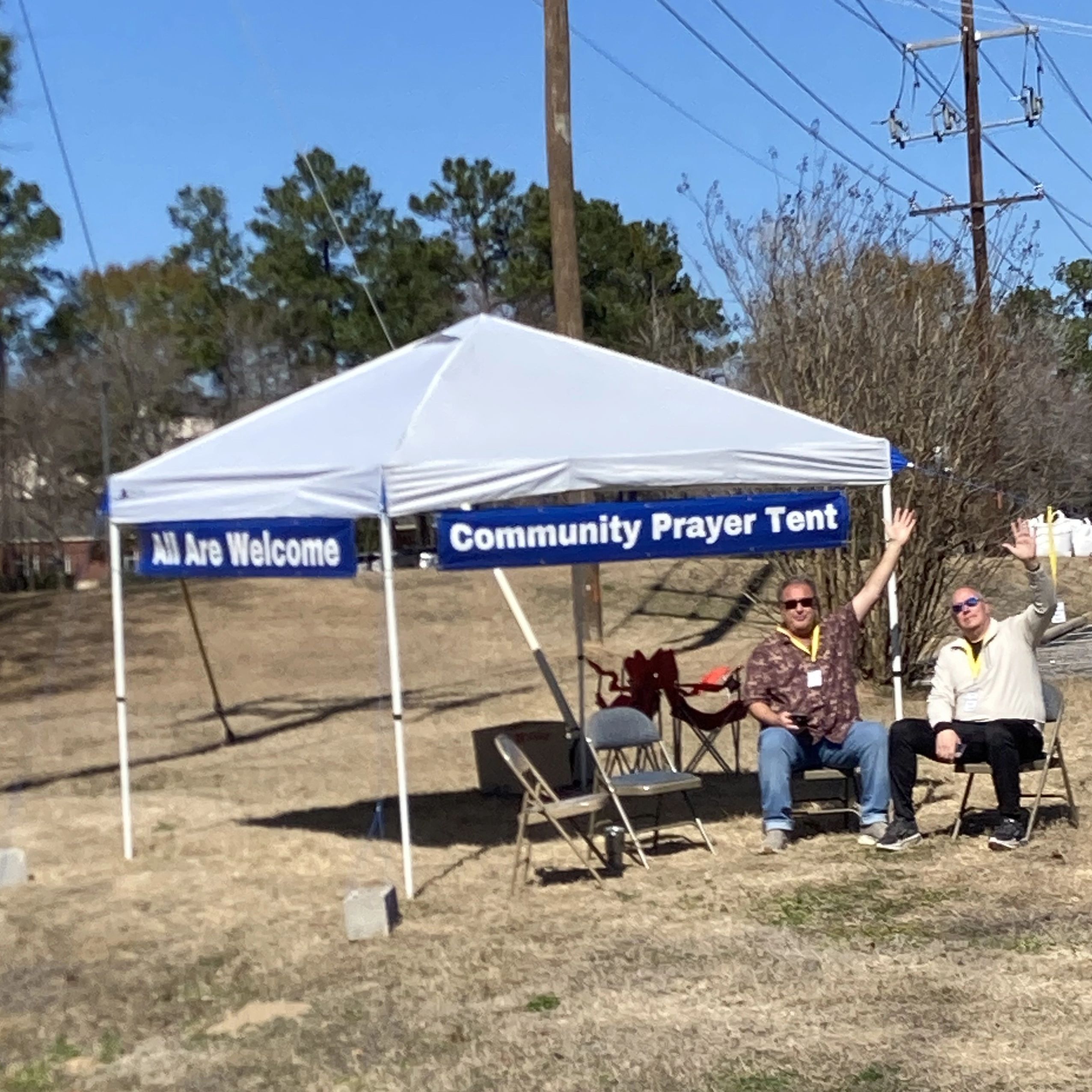 Two men seated under a white Community Prayer Tent on a sunny day.