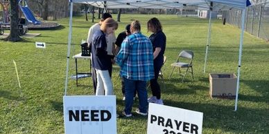 People gathered under a tent with signs asking for prayer support.