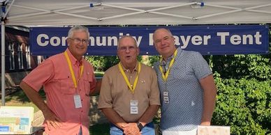 Three men stand under a Community Prayer Tent at an outdoor event.