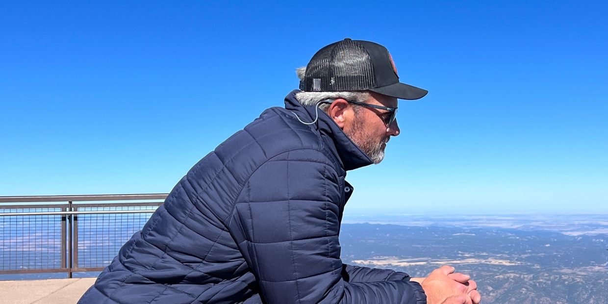 Man in a navy jacket and cap gazes thoughtfully at a vast mountain landscape.
