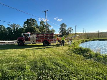 Fire truck and people standing near a pond in a rural area on a sunny day.