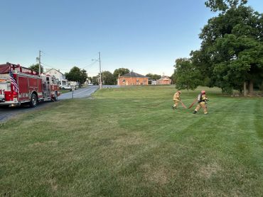 Two firefighters walk across a grassy field near a fire truck and residential houses.
