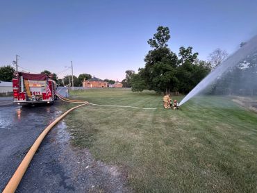 Firefighters using a hose to spray water from a fire truck onto a grassy area.