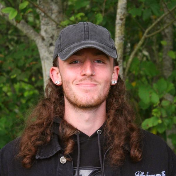 Young man with long curly hair wearing a cap and earrings outdoors.