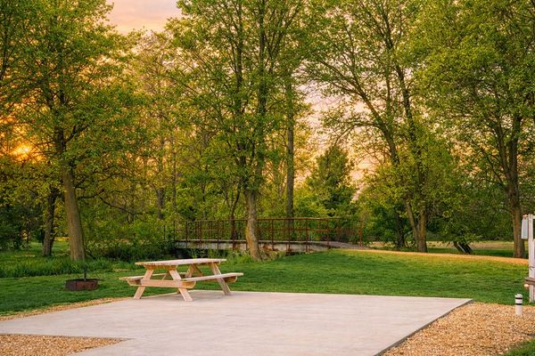 IMAGE OF PICNIC TABLE AT CAMPSPOT WITH WALKING BRIDGE IN BACKGROUND AND TREES WITH A SUNSET GLOW
