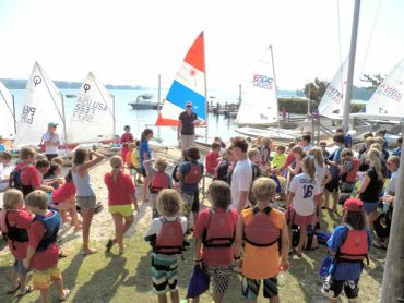 A group of children wearing life jackets gathers near sailboats by the water.