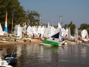 Small sailboats lined up on a beach with people preparing to sail.