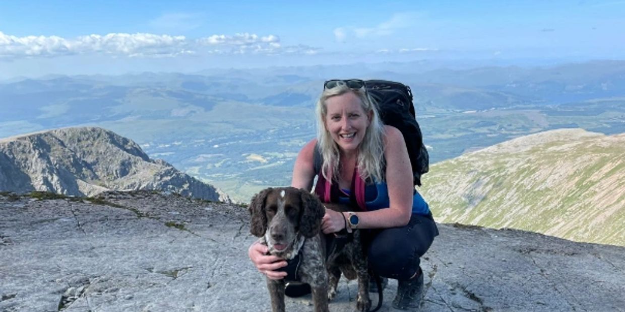 A woman with a backpack crouches and smiles beside her dog on a mountain summit.