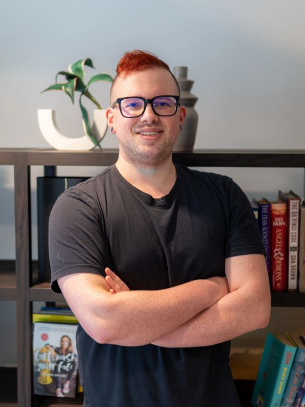 Smiling man with glasses and red hair stands confidently with arms crossed.