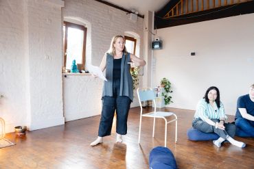 A woman stands and speaks in a bright room while others sit on the floor.