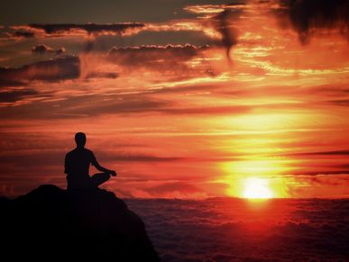 Person sitting on top of cliff, meditating with pretty clouds above them while watching the sunset.