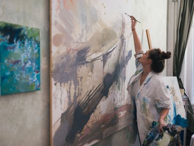 Female artist with hair in a bun is reaching tall with a brush to continue painting a large canvas.