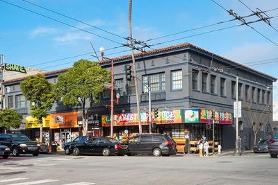 Colorful storefronts and vehicles line a sunny urban street corner.