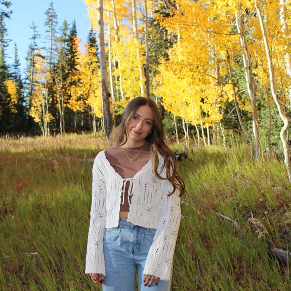 A woman stands in a grassy field with autumn yellow trees behind her.