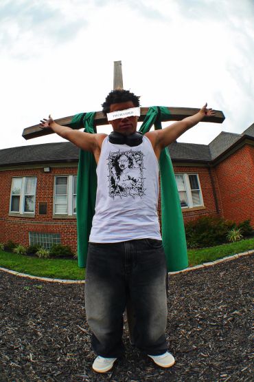 Person posing with arms outstretched in front of a cross outside a building.